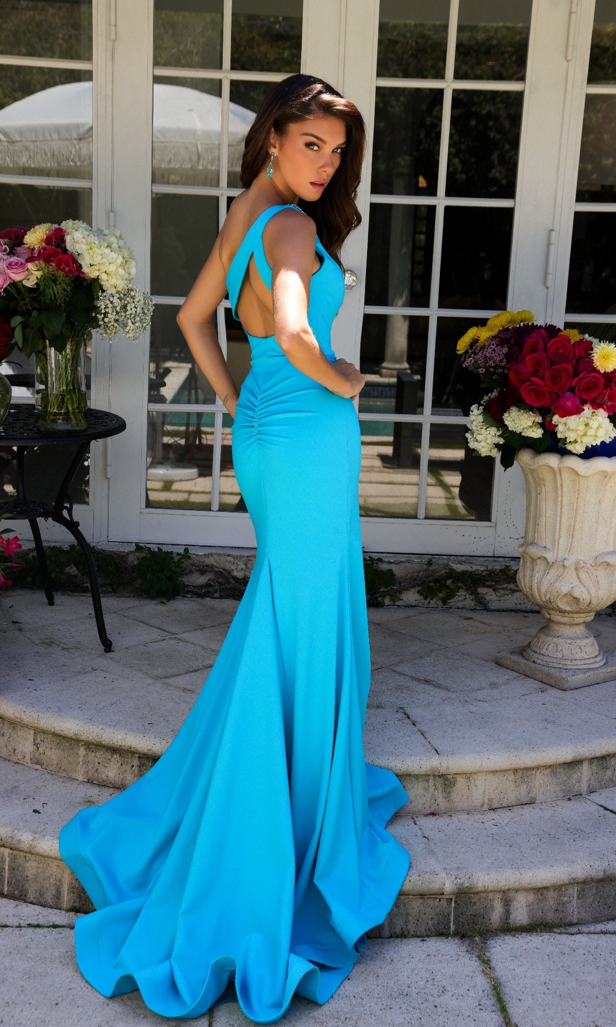 A woman stands outdoors in front of glass doors, surrounded by large floral arrangements, wearing the Ava Presley 40014 Keyhole Cut-Out One-Shoulder Prom Dress in bright blue.